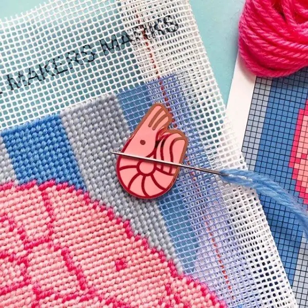 A foam shrimp illustrated needleminder holds a tapestry needle in place on top of a needlepoint canvas.