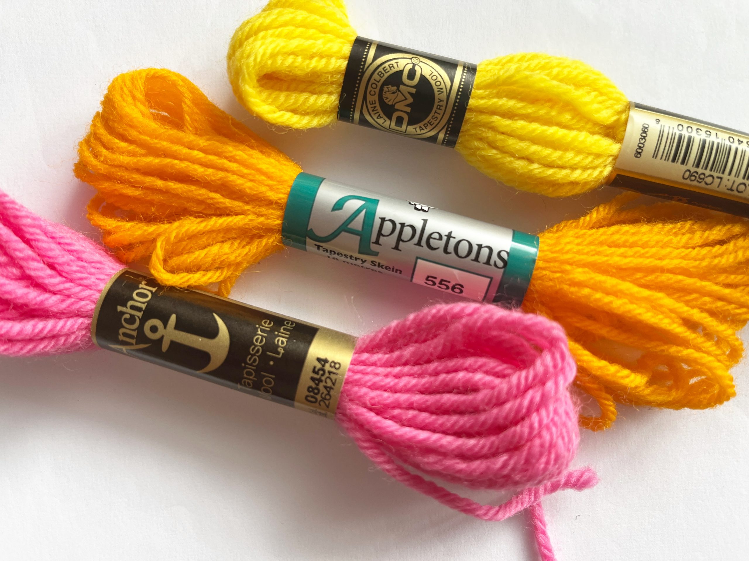 Three skeins of tapestry wool sit on a plain background. One skein is from Anchor, one from Appletons and one from DMC.